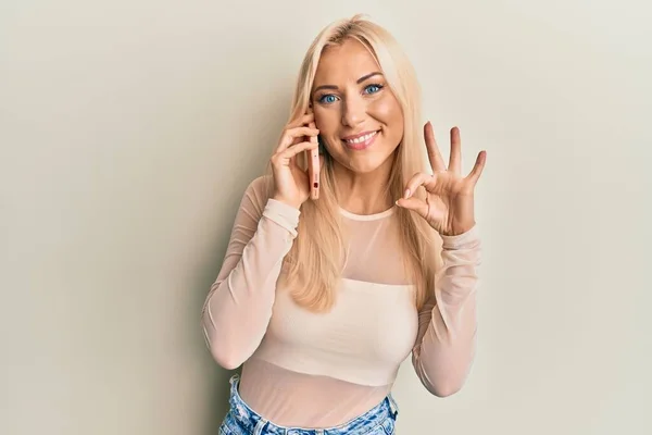 Młoda Blondynka Rozmawiając Smartfonie Robi Znak Palcami Uśmiechając Przyjazny Gest — Zdjęcie stockowe