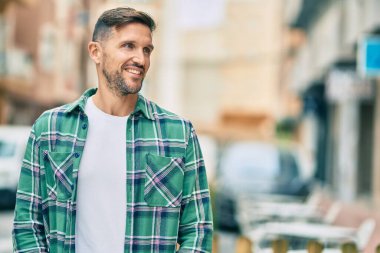 Genç beyaz adam gülümseyerek şehirde mutlu mesut duruyor..
