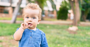 Üzgün küçük çocuk, güneşli bir günde parkta diş etlerine dokunduğu için parmaklarını dudaklarına koyuyor. Güzel sarı saçlı erkek bebek yeni bebek dişleri için acı çekiyor.