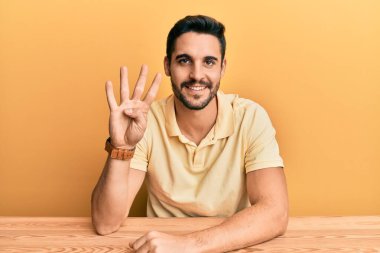 Latin asıllı genç adam, gündelik giysiler içinde masada oturuyor ve dört numaralı parmaklarıyla işaret ederken kendinden emin ve mutlu gülümsüyor.. 