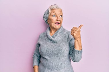 Kıdemli gri saçlı kadın yün kazak ve kış şapkası takıyor. Gülümsüyor, gülümsüyor ve baş parmağıyla yan tarafa bakıyor.. 