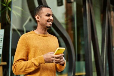 Genç Afrikalı Amerikalı adam şehirde akıllı telefon kullanarak mutlu bir şekilde gülümsüyor..