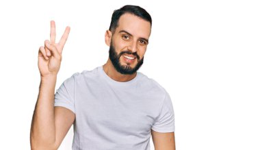 Sakallı, günlük beyaz tişörtlü, gülümseyen ve parmaklarını zafer işareti yaparken gösteren genç bir adam. İki numara.. 