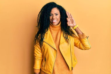 Orta yaşlı Afro-Amerikalı kadın yün kazak ve deri ceket giyip gülümsüyor ve el ve parmaklarıyla imza atıyor. başarılı ifade. 
