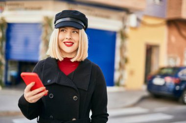 Genç sarışın kadın şehirde akıllı telefon kullanarak mutlu bir şekilde gülümsüyor..