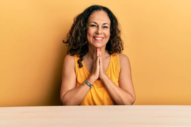 Orta yaşlı, güzel bir kadın, gündelik giysiler içinde masada oturmuş, elleriyle dua ediyor, af diliyor, kendinden emin bir şekilde gülümsüyor.. 