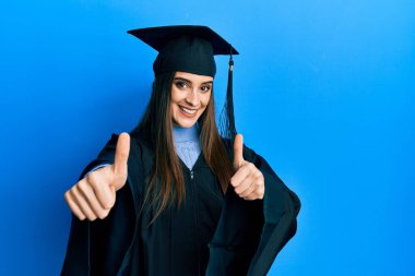 Mezuniyet şapkası ve tören cübbesi takan güzel esmer bir kadın elleriyle olumlu bir jest yapmayı onaylıyor, gülümsüyor ve başarı için seviniyor. kazanan jest. 