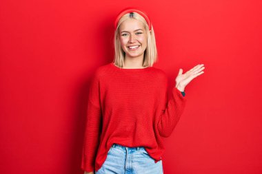 Güzel, kırmızı süveter giyen, neşeli bir şekilde gülümseyen ve avuç içi kameraya bakan sarışın bir kadın.. 