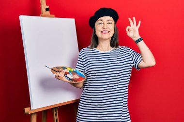 Orta yaşlı, İspanyol bir kadın boya sehpası ile resim çiziyor gülümsüyor ve el ve parmaklarıyla imza atıyor. başarılı ifade. 