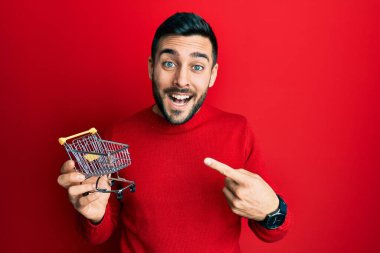 Genç İspanyol adam elinde küçük bir süpermarket arabası tutarken mutlu bir şekilde el ve parmak işareti yapıyor. 