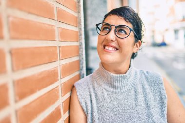 Genç ve cüsseli bir kadın gülümsüyor. Şehrin duvarına yaslanmaktan mutlu..