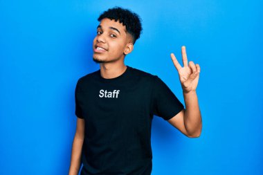 Genç Afro-Amerikalı, personel tişörtü giymiş gülümseyen ve parmaklarını zafer işareti yaparken gösteren kameraya bakan bir adam. İki numara.. 