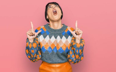 Gündelik kıyafetler giyen genç İspanyol kadın şaşırmış ve şaşırmış bir şekilde yukarı bakıyor ve parmaklarıyla işaret ediyor ve kollarını kaldırıyor.. 