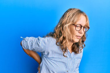 Orta yaşlı sarışın kadın gündelik kıyafetler giyiyor ve gözlük takıyor sırt ağrısı çekiyor, elleriyle dokunuyor, kas ağrısı çekiyor. 