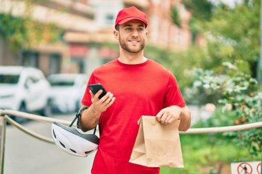 Beyaz atlı genç teslimatçı akıllı telefon kullanıyor ve şehirde kağıt torba taşıyor..