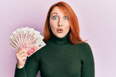 Güzel kızıl saçlı kadın elinde 20 İsrail şekeri banknotları tutuyor. Korkmuş ve şaşırmış bir şekilde ağzı açık, inançsız bir yüz. 