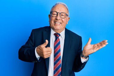 Beyaz üst düzey bir adam iş elbisesi ve kravat takıyor. Avuç içi gösteriyor ve baş parmağıyla iyi bir hareket yapıyor. Mutlu ve neşeli gülümsüyor. 