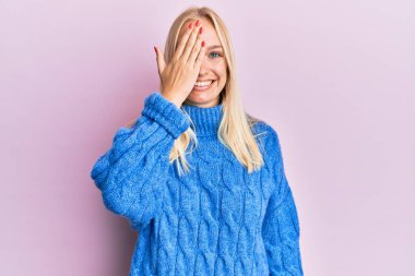 Kışın yün kazak giyen genç sarışın kız bir gözünü eliyle kapatıyor, yüzünde kendine güvenen bir gülümseme ve sürpriz bir duygu var.. 