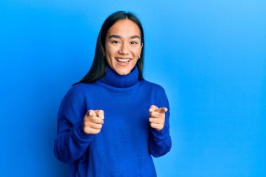 Kış süveteri giyen genç Asyalı kadın mutlu ve komik yüzüyle kamerayı işaret ediyor. iyi enerji ve titreşimler. 