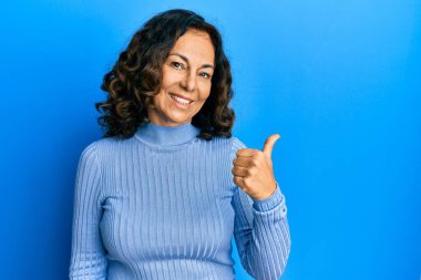 Orta yaşlı İspanyol kadın, günlük kıyafetler giyiyor, gülümsüyor, gülümsüyor ve başparmağıyla yan tarafa bakıyor.. 