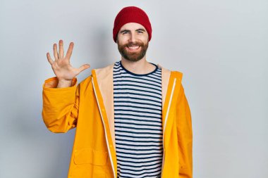 Sakallı, sarı yağmurluk giyen beyaz bir adam beş numaralı parmaklarıyla işaret ederken kendinden emin ve mutlu bir şekilde gülümsüyor.. 