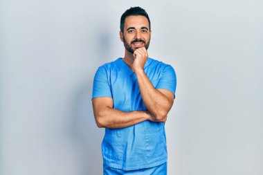 Sakallı, mavi erkek hemşire üniforması giyen yakışıklı, gülümseyen kolları çapraz ve çenesi kalkık bir adam. Olumlu düşünmek. 