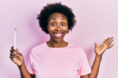 Genç Afrikalı Amerikalı kadın elinde elektrikli diş fırçasıyla zaferi kutluyor mutlu bir gülümsemeyle ve ellerini kaldırarak kazanan bir ifadeyle. 