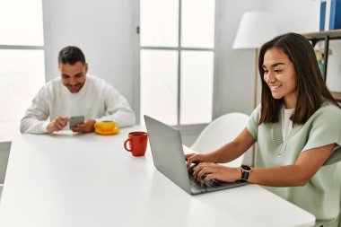 Laptop ve akıllı telefon kullanan genç latin çift evde kahve içiyor..
