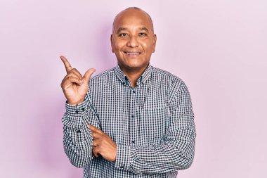 Orta yaşlı, sıradan kıyafetler giyen, yüzünde kocaman bir gülümseme olan, eli ve parmağı kameraya bakan Latin bir adam.. 