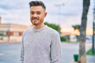 Genç beyaz adam gülümseyerek şehirde mutlu mesut duruyor..