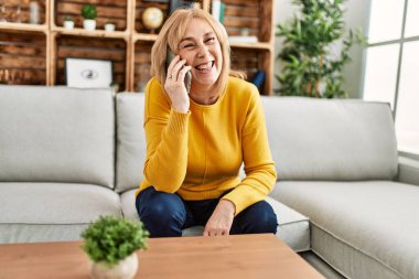 Orta yaşlı sarışın kadın akıllı telefonda konuşuyor. Evdeki kanepede oturuyor..