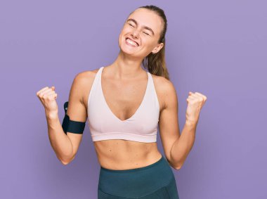 Spor giyim ve kol bandı takan güzel sarışın kadın çok mutlu ve heyecanlı bir şekilde kolları havada, gülümseyen ve başarı için bağıran bir jest yapıyor. kutlama konsepti. 