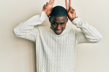 Genç Afro-Amerikan adam günlük kıyafetler giyiyor komik ve çılgın poz veriyor parmakları tavşan kulağı gibi, neşeli gülümsüyor 