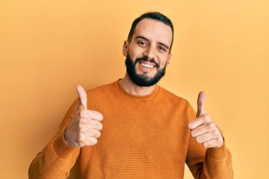 Sakallı genç adam, günlük kışlık süveter giyer, başarı işareti yapar, elleriyle olumlu jest yapar, gülümser ve mutlu olur. Neşeli ifade ve kazanan jest. 