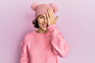 Genç esmer, sevimli yün şapkalı bir kadın bir gözünü eliyle kapatıyor, yüzünde kendine güvenen bir gülümseme ve şaşırtıcı bir duygu var.. 