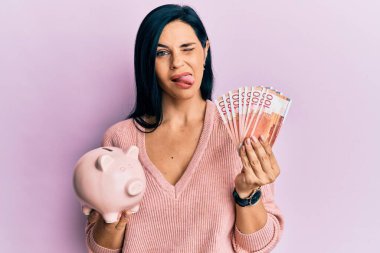 Norveç kronu banknotları ve domuz kumbarası tutan beyaz bir kadın komik bir ifadeyle dilini dışarı çıkarıyor.. 