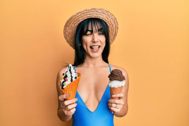 Kahküllü, mayo giyen, elinde iki dondurma külahı olan genç esmer kadın kameraya seksi bir ifadeyle bakıyor. Neşeli ve mutlu bir yüzü var.. 