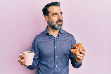 Orta yaşlı, sakallı, kır saçlı, kahve içen, pasta yiyen, gülümseyen ve kafasını çeviren bir adam.. 