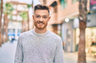 Genç beyaz adam gülümseyerek şehirde mutlu mesut duruyor..