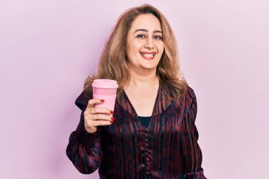 Orta yaşlı, beyaz bir kadın kahvesini içerken pozitif ve mutlu görünüyor. Kendine güvenen bir gülümsemeyle dişlerini gösteriyor. 