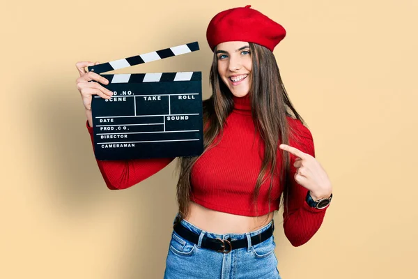 Young brunette teenager holding video film clapboard smiling happy pointing with hand and finger