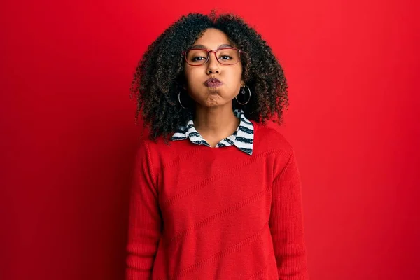 Beautiful african american woman with afro hair wearing sweater and glasses puffing cheeks with funny face. mouth inflated with air, crazy expression.