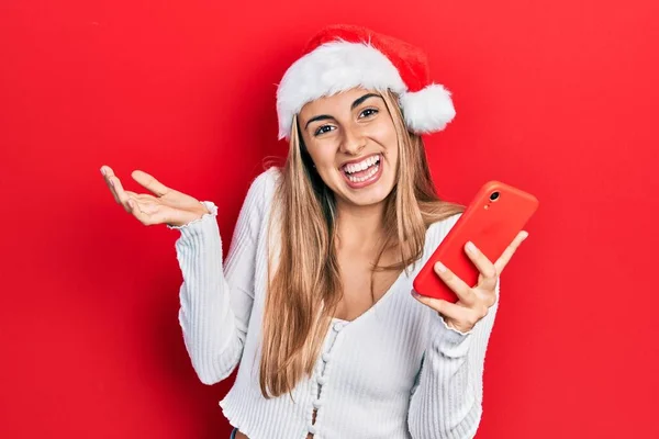 Όμορφη Ισπανόφωνη Γυναίκα Φορώντας Χριστουγεννιάτικο Καπέλο Κρατώντας Smartphone Γιορτάζει Επίτευγμα — Φωτογραφία Αρχείου