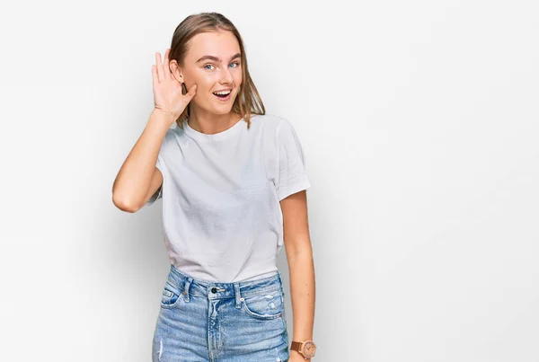 噂やゴシップに耳を傾ける耳の上に手で笑みを浮かべてカジュアルな白いTシャツを着て美しい若いブロンドの女性 聴覚障害の概念 — ストック写真