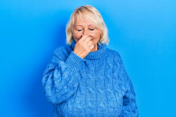 Middle age blonde woman wearing casual clothes smelling something stinky and disgusting, intolerable smell, holding breath with fingers on nose. bad smell