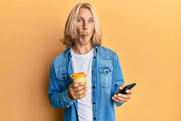 Caucasian young man with long hair using smartphone and drinking a cup of coffee making fish face with mouth and squinting eyes, crazy and comical.