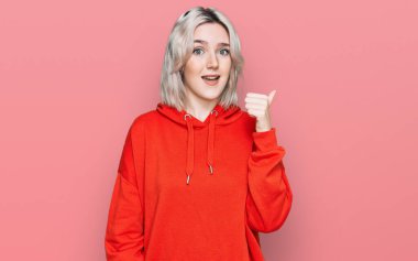 Gündelik kıyafetler giyen, gülen yüzlü genç sarışın kız başparmağıyla yan tarafa bakıyor ve işaret ediyor.. 