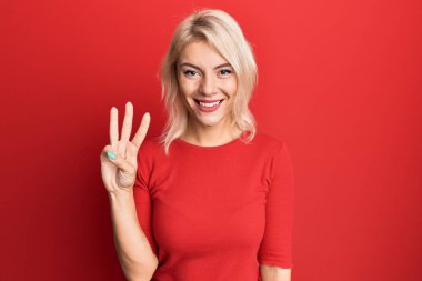 Gündelik kıyafetler giyen genç sarışın kız üç numarayı gösterip işaret ederken kendinden emin ve mutlu gülümsüyor.. 