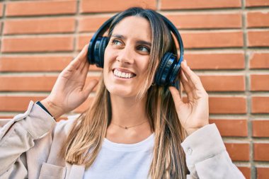 Genç beyaz kadın gülümseyerek şehirde kulaklık takarak müzik dinliyor..