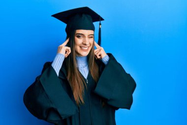 Mezuniyet şapkası ve tören cübbesi takan güzel esmer bir kadın yüksek sesli müzik sesi için kulaklarını parmaklarıyla kapatıyor. sağır kavramı. 